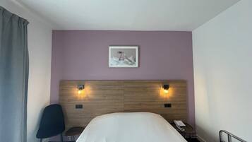 Habitación de hotel con una cama, una silla, una mesita, un teléfono y un cuadro en la pared.