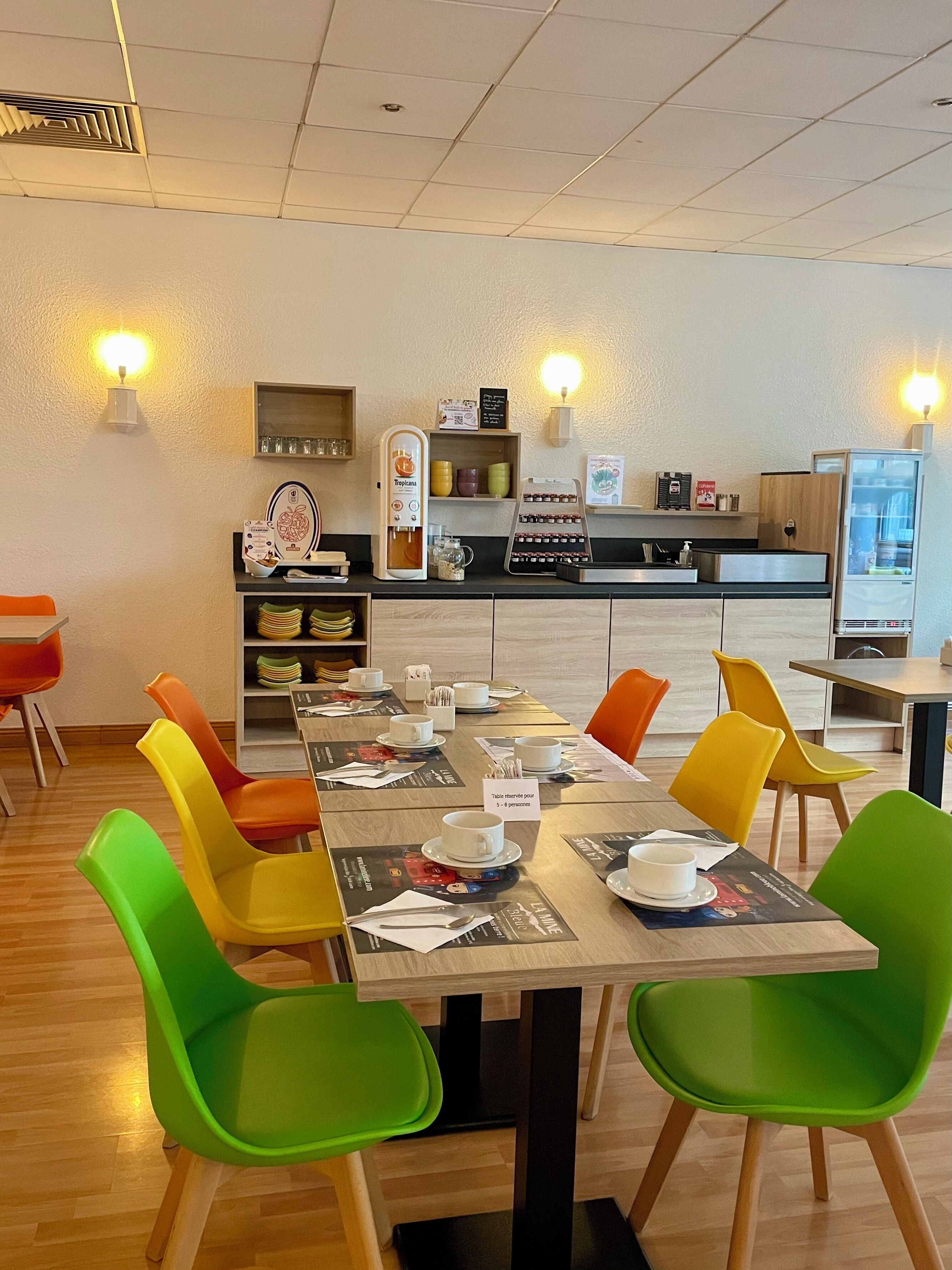 A dining area with a long table set for 5-6 people, colorful chairs, and a coffee machine.