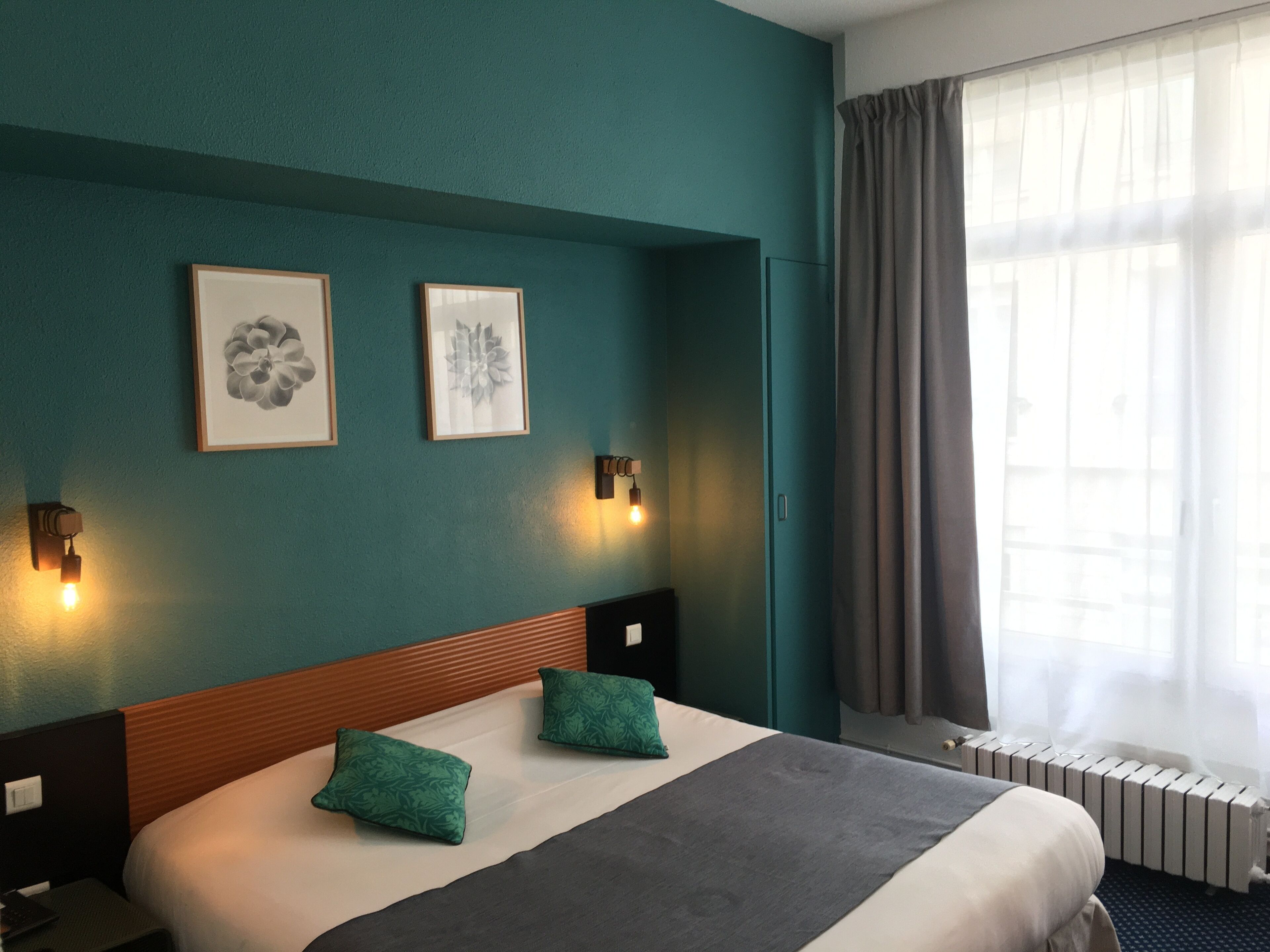A hotel room with a large bed, two pillows, a headboard, wall-mounted lights, and two framed floral artworks.