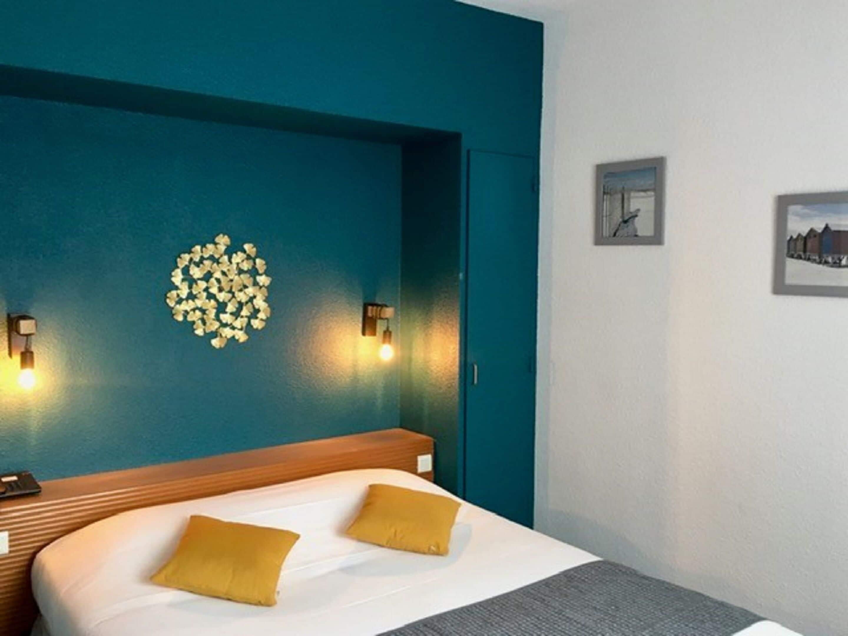 A hotel room with a teal accent wall, a bed with two yellow pillows, a wooden headboard, and two wall-mounted lights.
