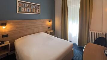 A hotel room with a large bed, a headboard with built-in lighting, a small bedside table, a chair, a window with curtains, and a framed picture on the wall.