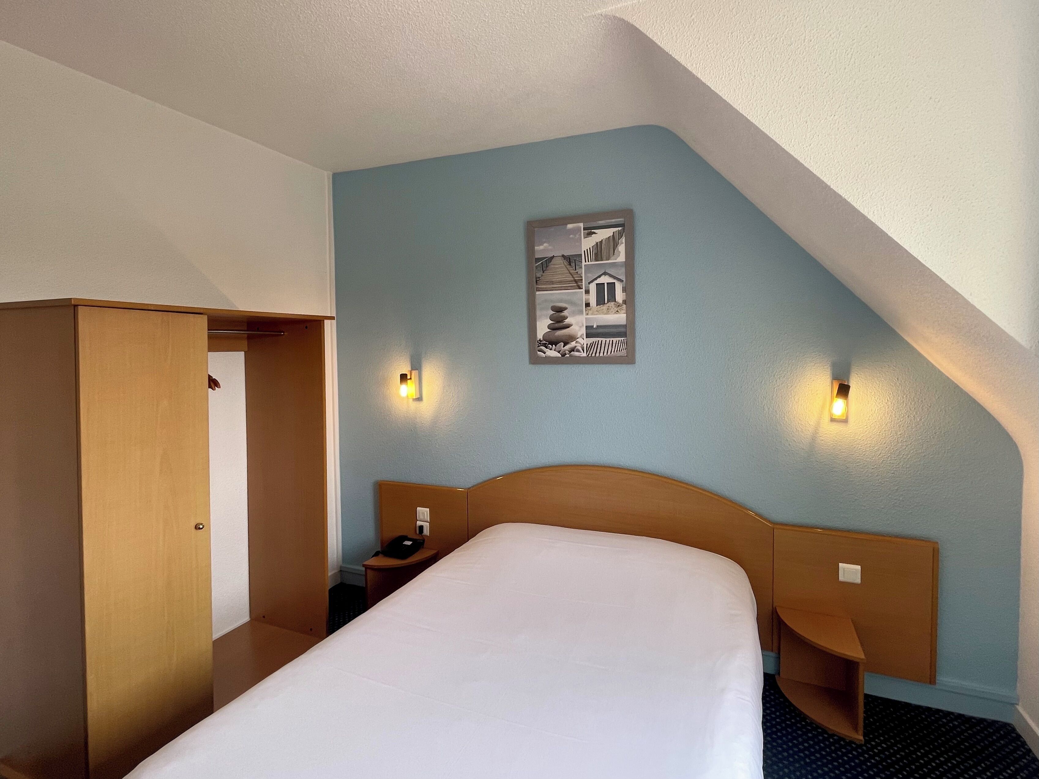 Et hotelværelse med seng, et træskab, et natbord og en lampe på væggen.