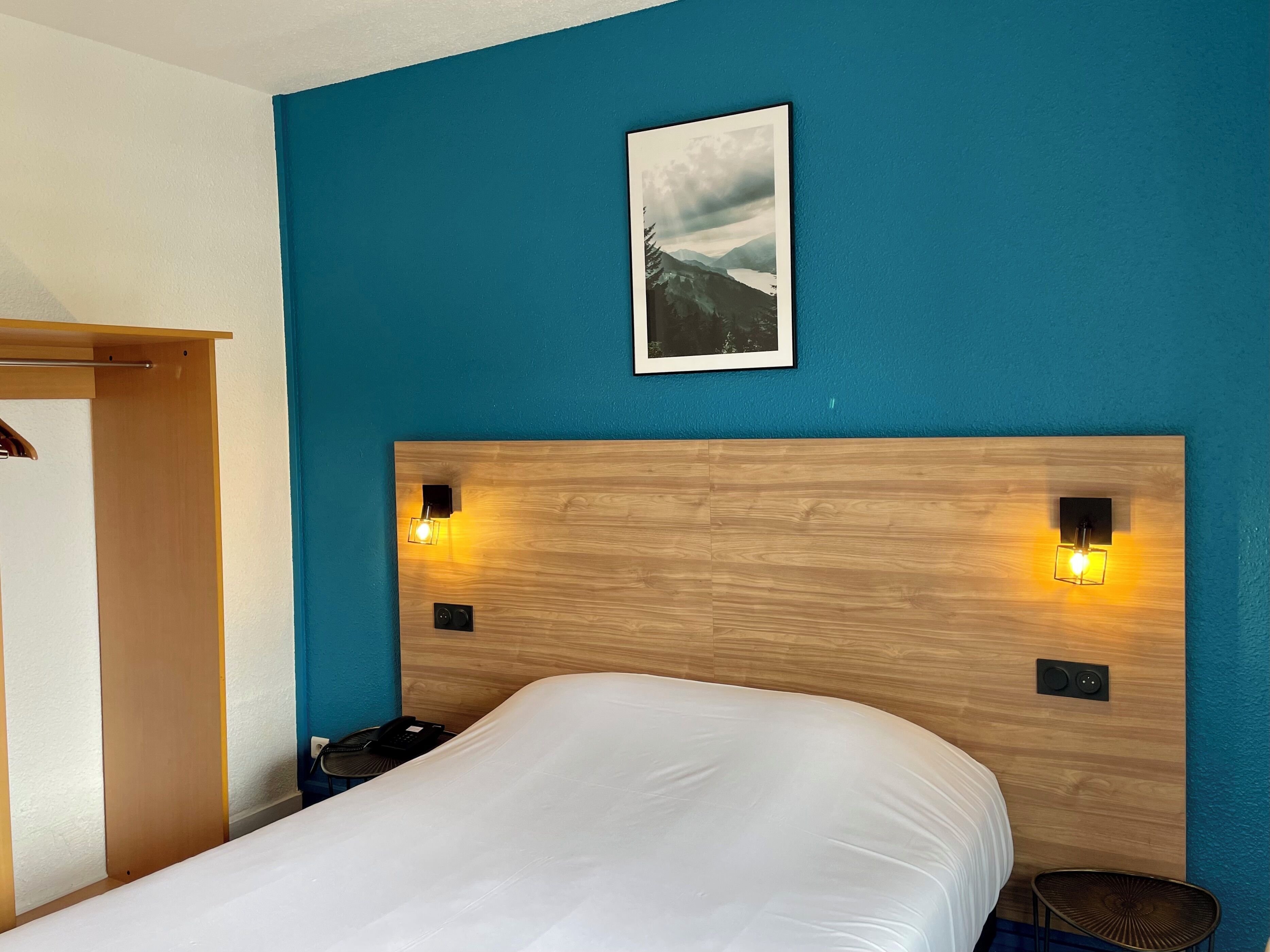 Habitación de hotel con cabecera de madera, dos lámparas de pared, cama con ropa blanca, mesita de noche pequeña, una silla e imagen enmarcadas en la pared.