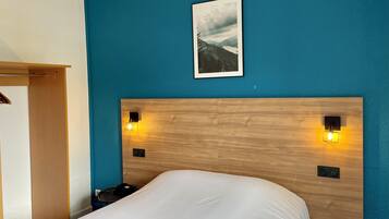 Habitación de hotel con cabecera de madera, dos lámparas de pared, cama con ropa blanca, mesita de noche pequeña, una silla e imagen enmarcadas en la pared.