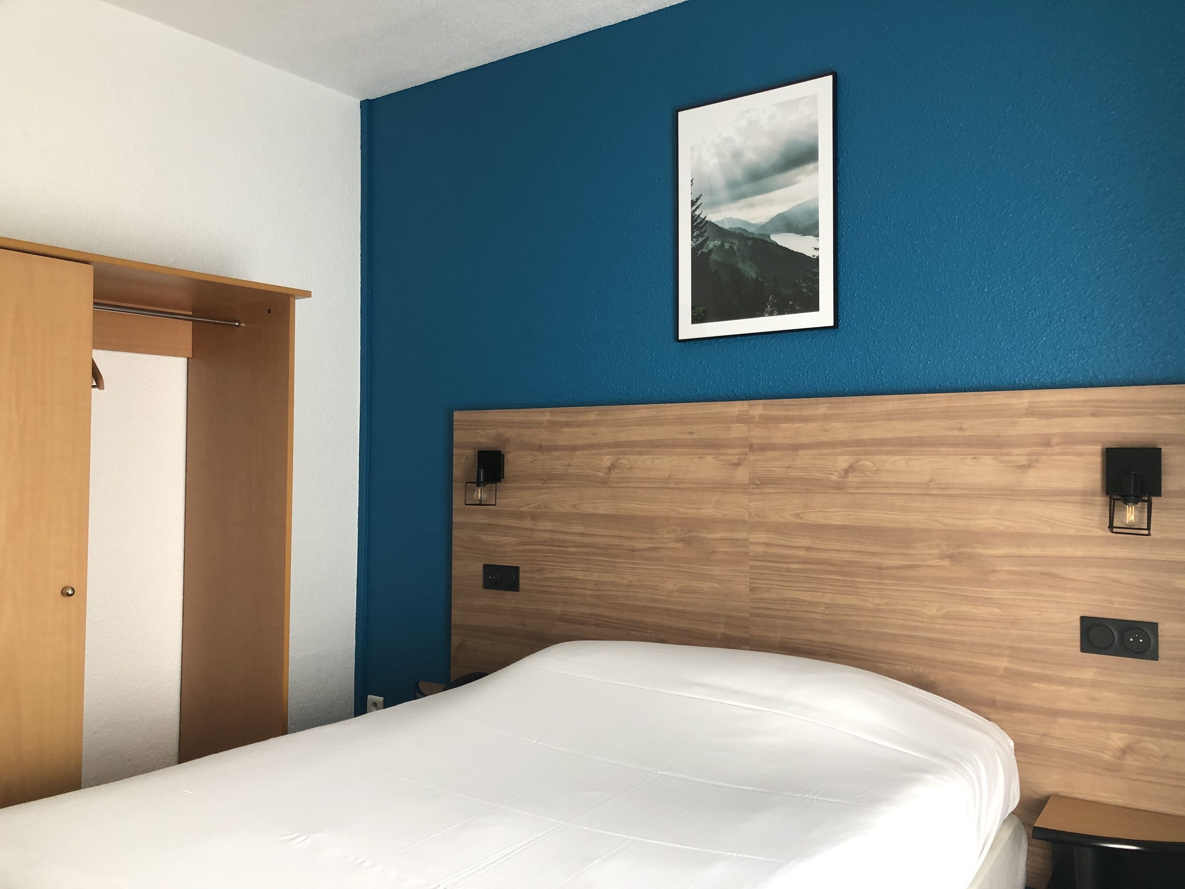 A bedroom with a wooden headboard, a white mattress, a framed picture on the wall, and a wooden door.