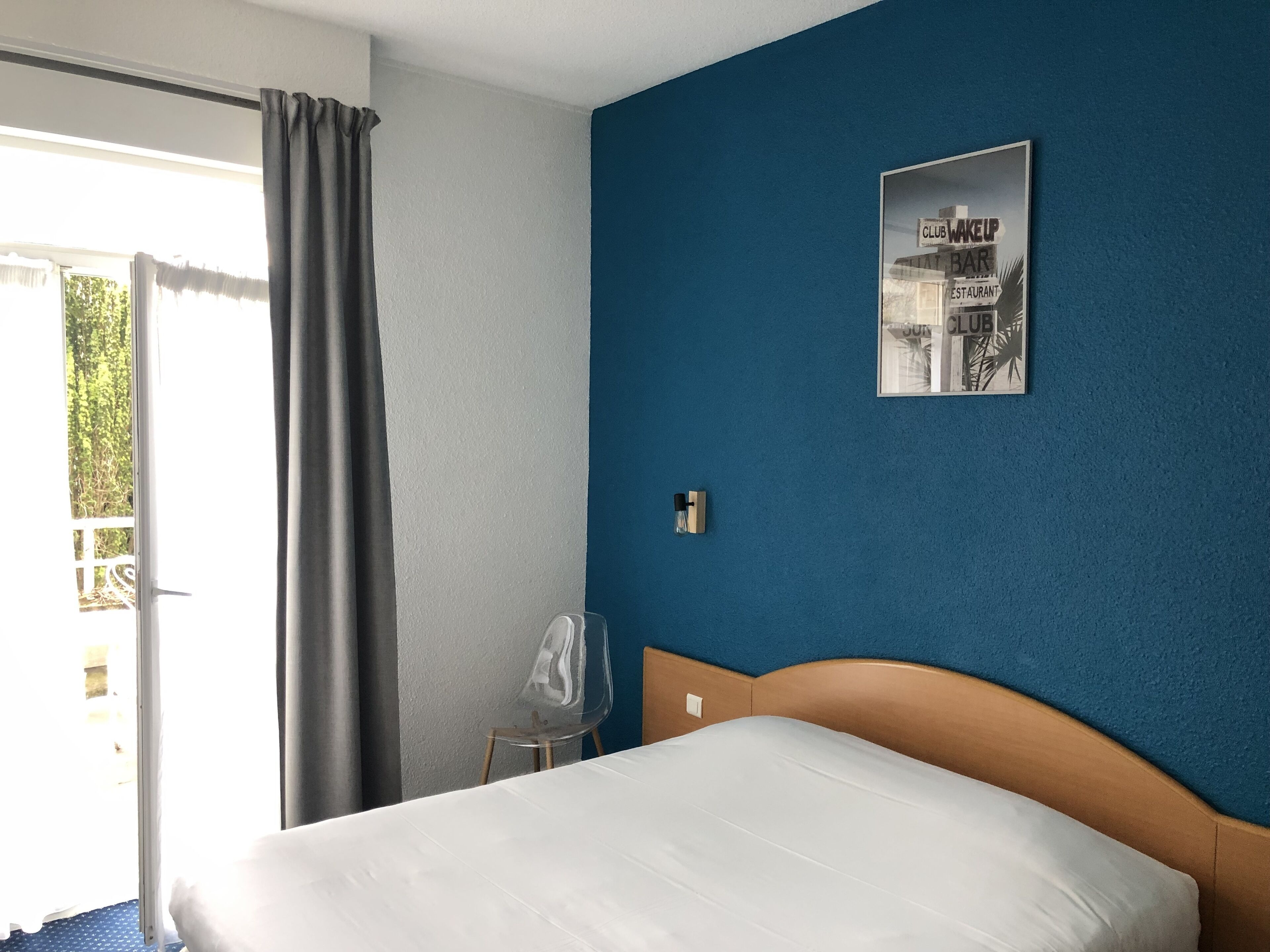 A hotel room with a bed, a chair, a window with curtains, and a wall with a poster.