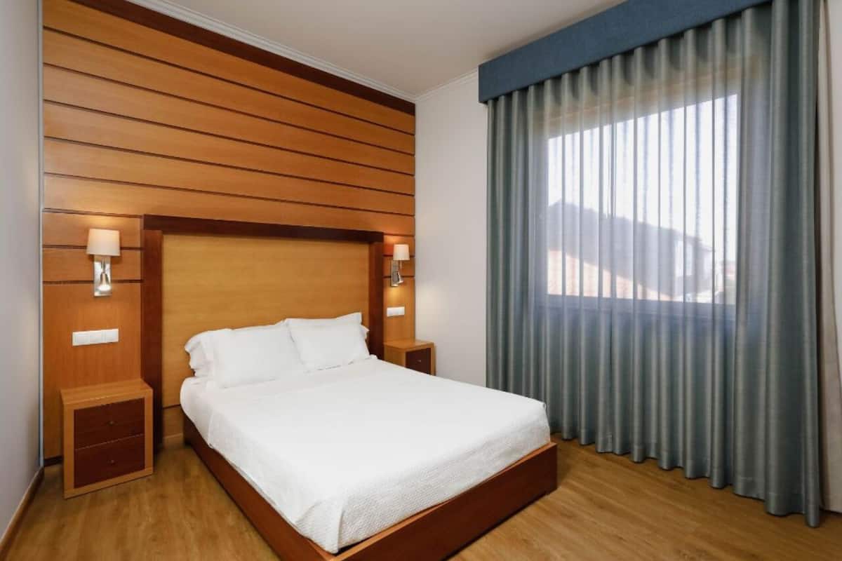 A hotel room with a wooden headboard, a large bed, two bedside tables with lamps, and a view through a window with curtains.