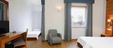 A hotel room with a wooden desk, a grey armchair, a bed, and a window with curtains.