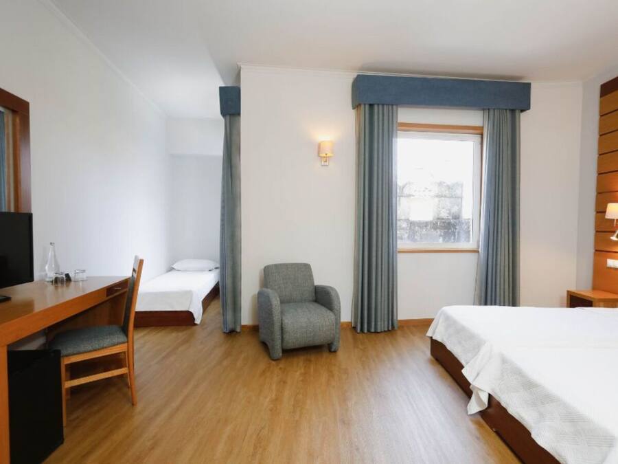 Ein Hotelzimmer mit einem Holztisch, einem grauen Sessel, einem Bett und einem Fenster mit Vorhängen.