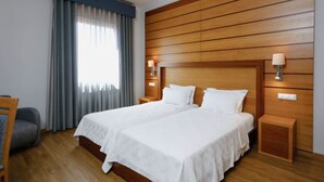 A double bed with white linens, a wooden headboard, two bedside tables with lamps, a grey armchair, and a window with grey curtains. - Montebelo Palácio dos Melos Viseu Historic Hotel (Viseu)