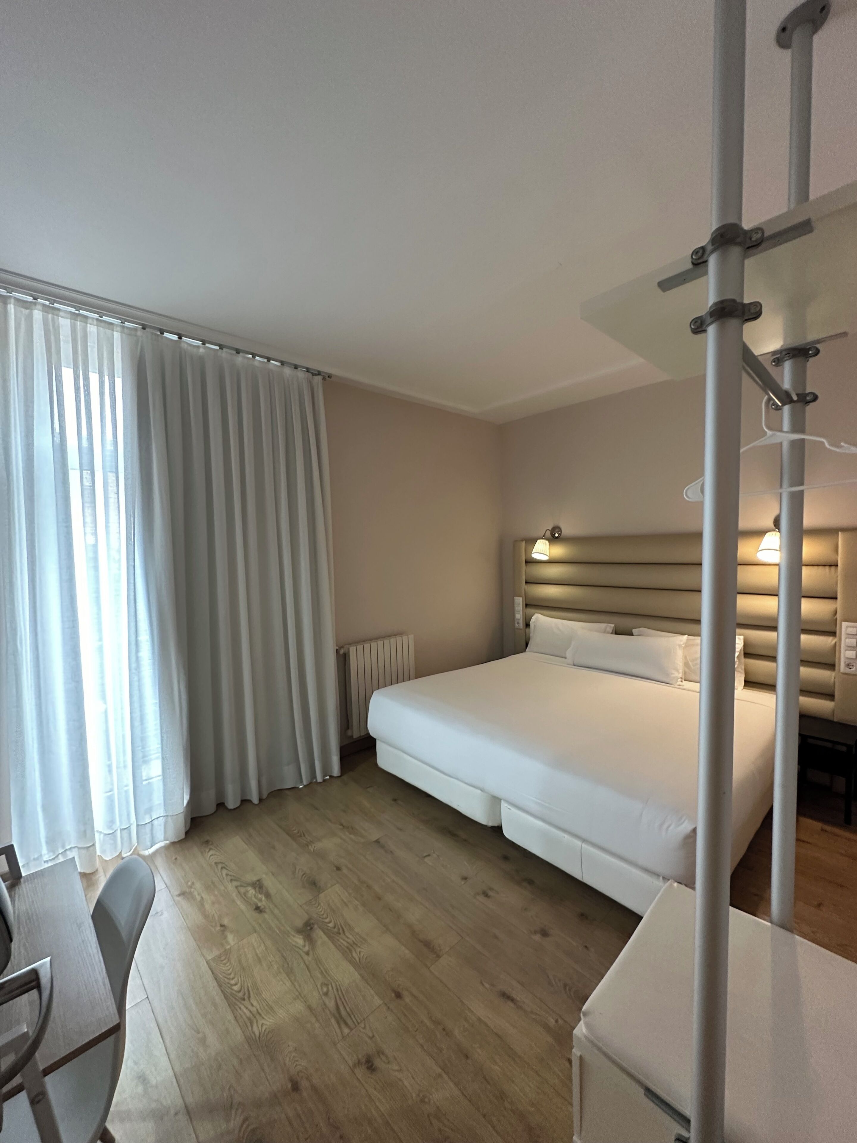 A hotel room with a bed, a desk, a chair, a radiator, and curtains.