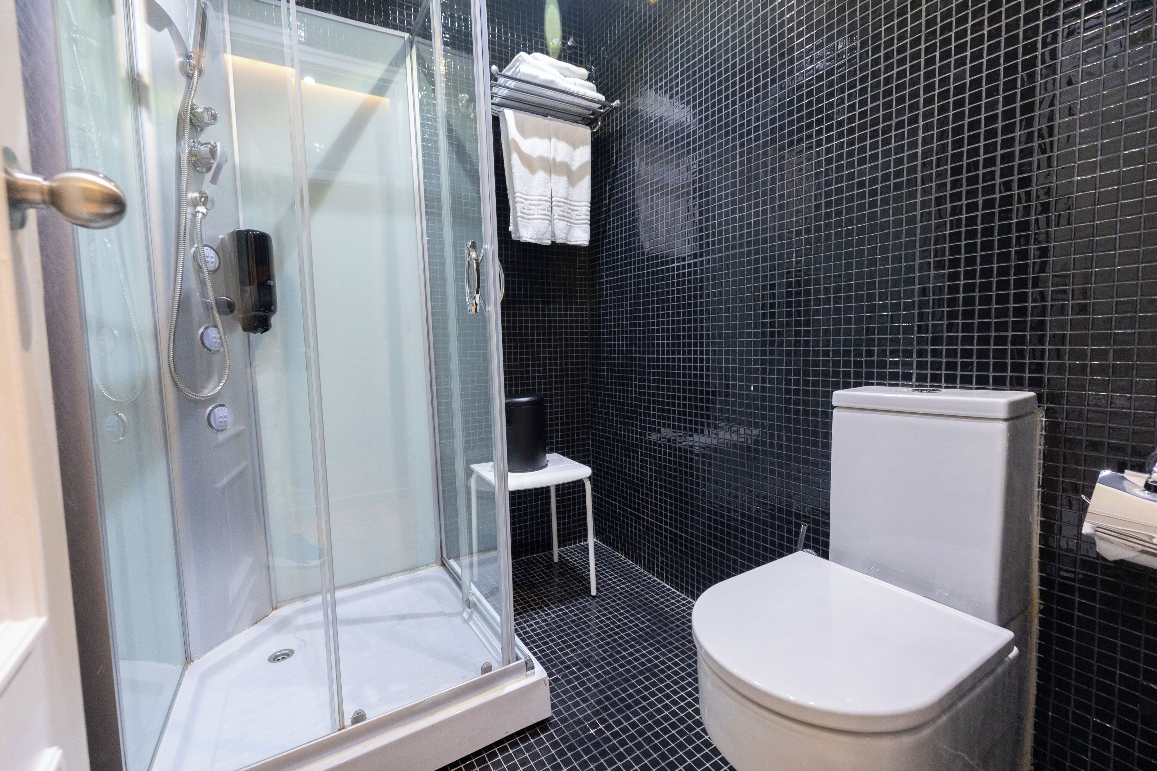 Un baño con ducha de vidrio, inodoro blanco y un taburete pequeño blanco.