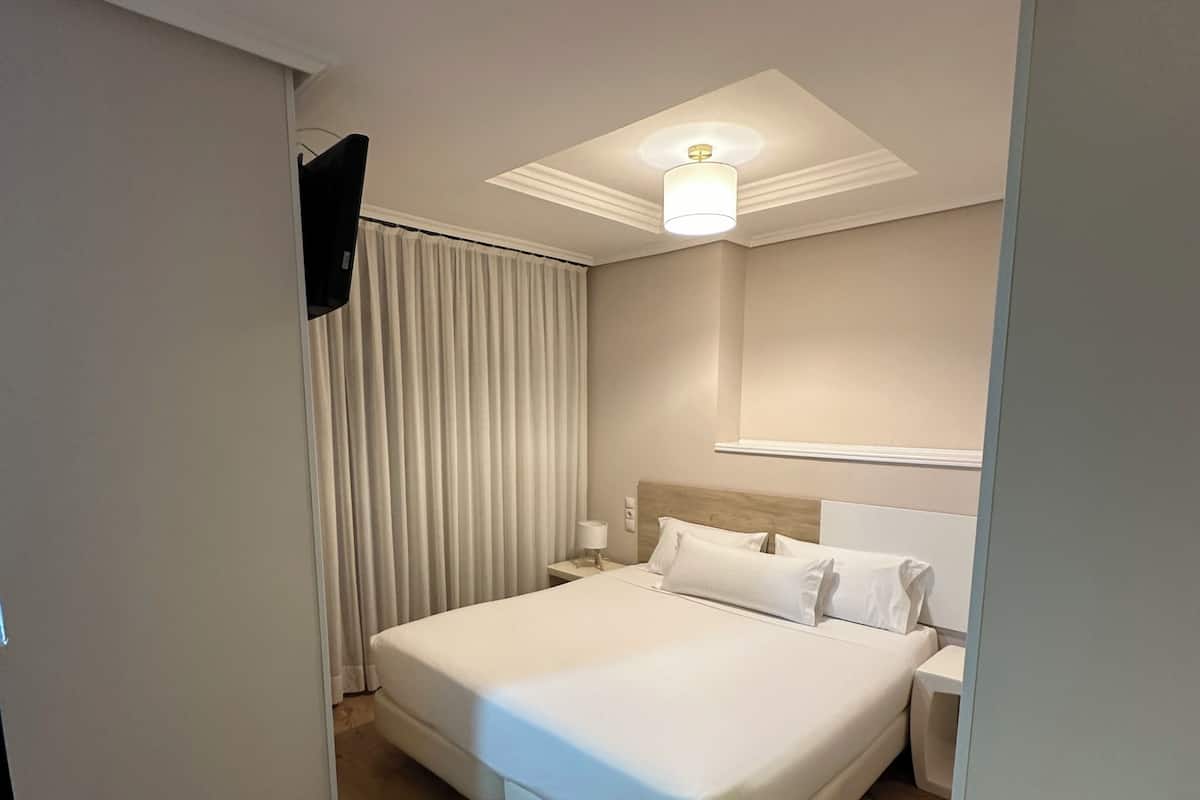 A hotel room with a large bed, a bedside table, a flat-screen TV, and a ceiling light fixture.