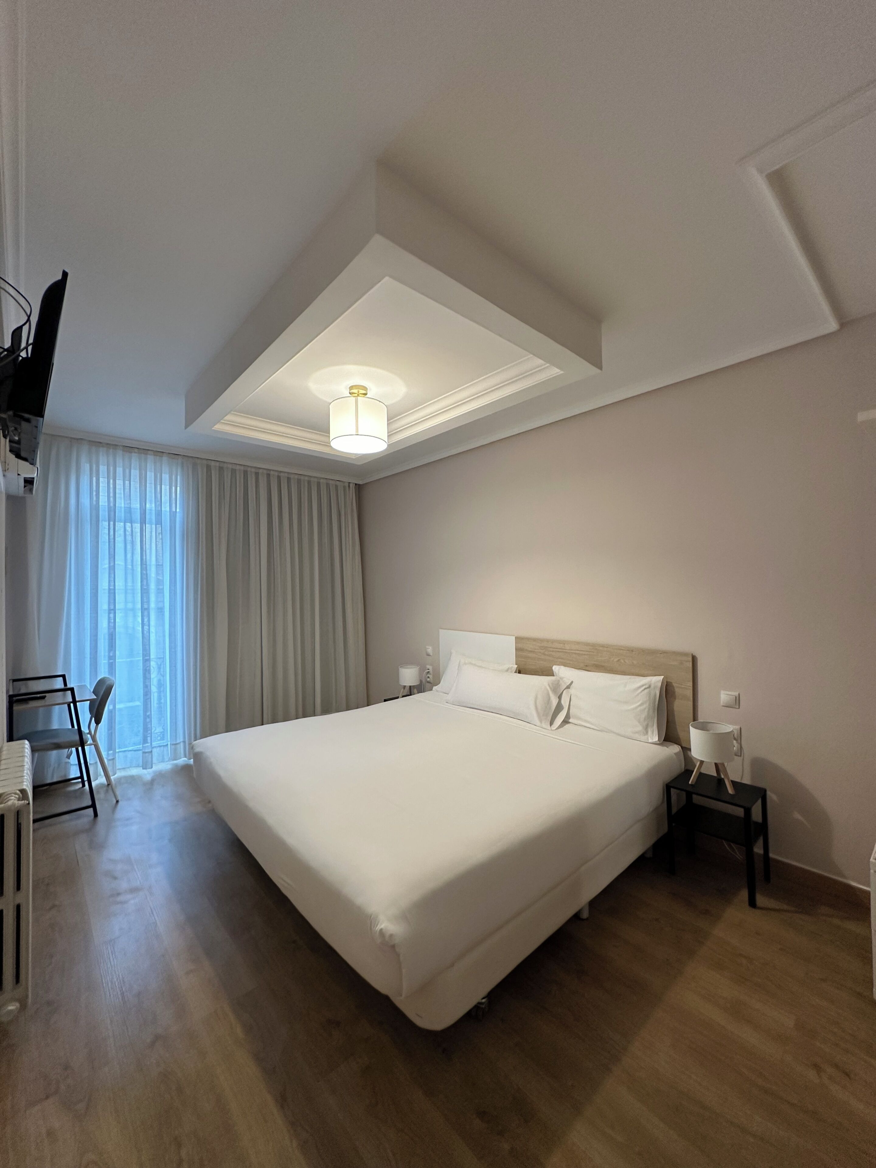Una camera d'albergo con un letto grande, una scrivania con una sedia, una TV a muro e un lampadario.