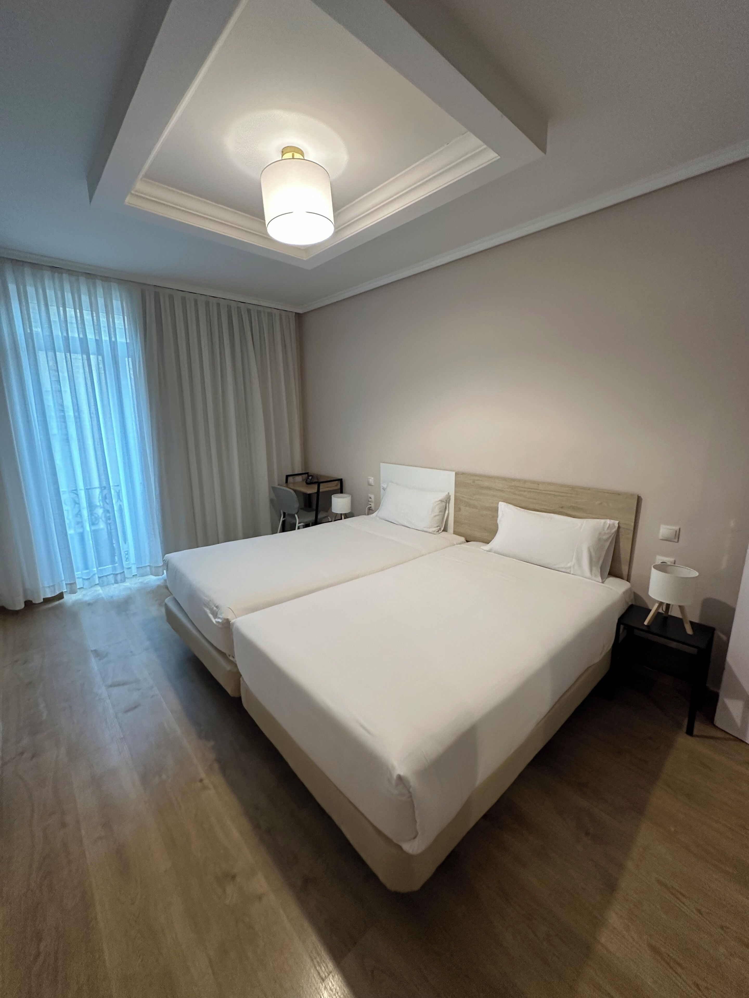 A hotel room with a large bed, a ceiling light, a bedside table, and a window with curtains.