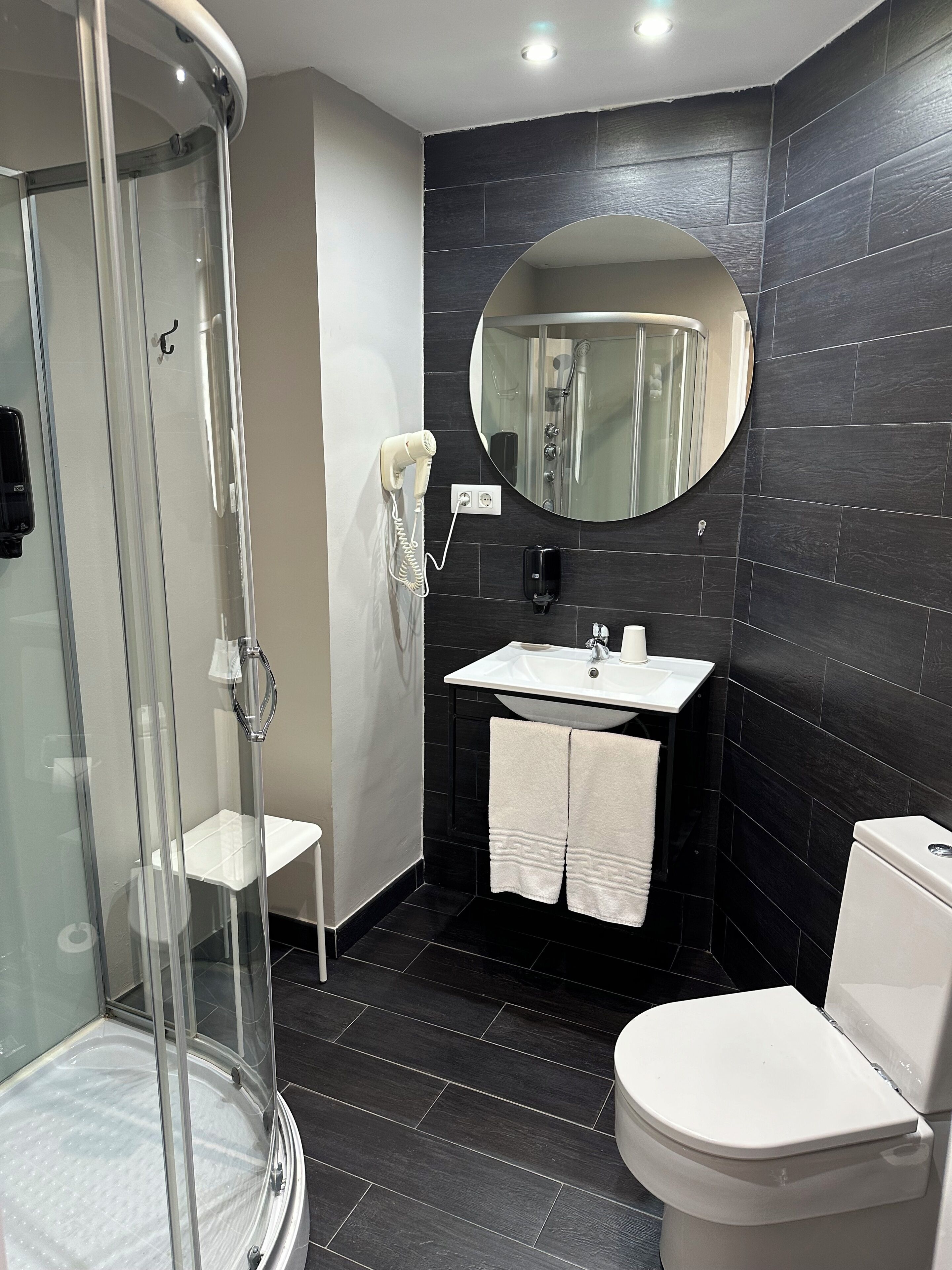 A modern bathroom with a glass-enclosed shower, a white sink, a round mirror, and a toilet.