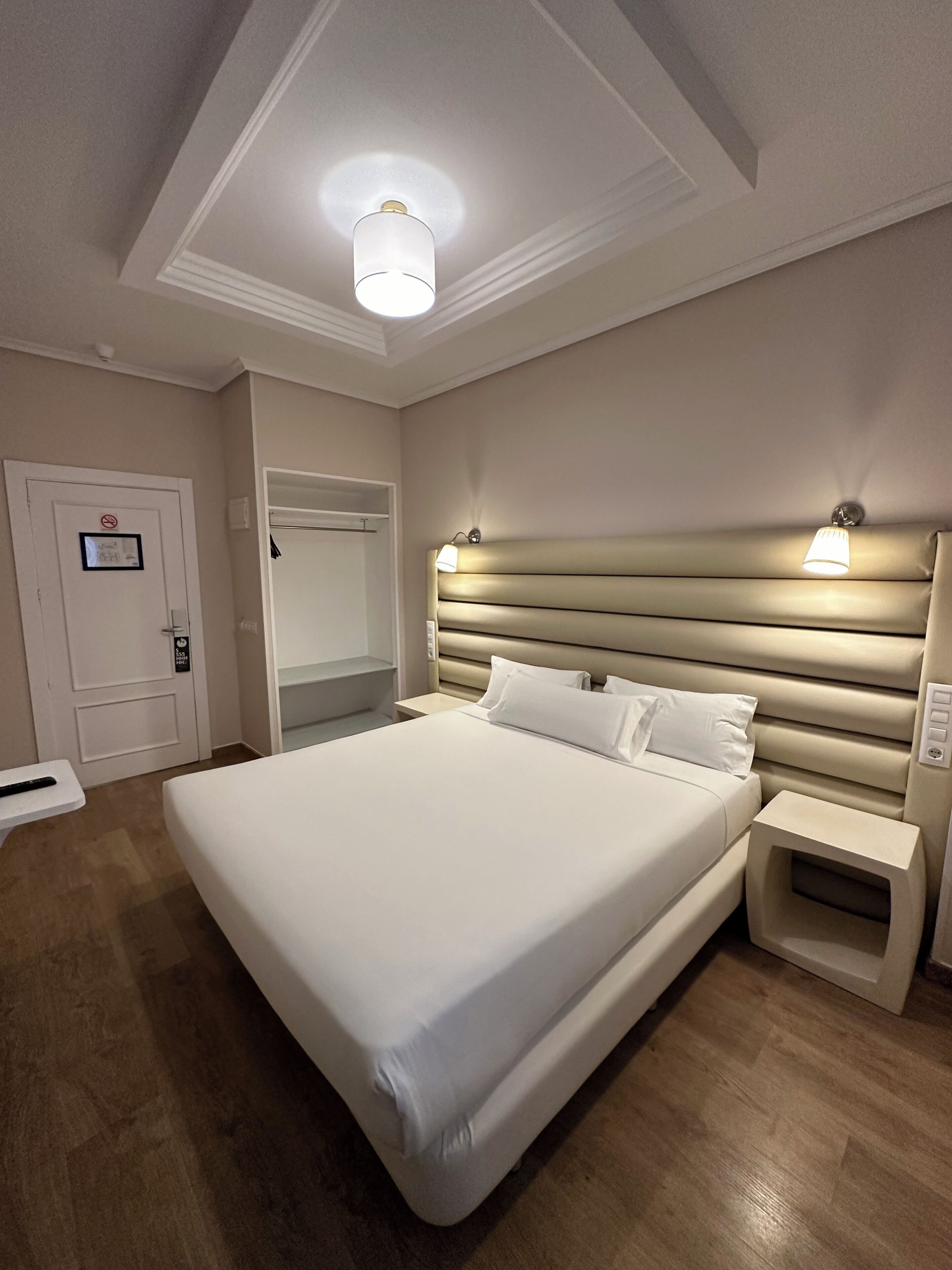 Habitación de hotel con una cama grande, mesitas de noche, una lámpara de techo y una puerta con un cartel.