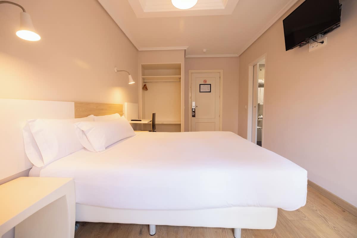 A hotel room with a bed, bedside table, lamp, and a television mounted on the wall.