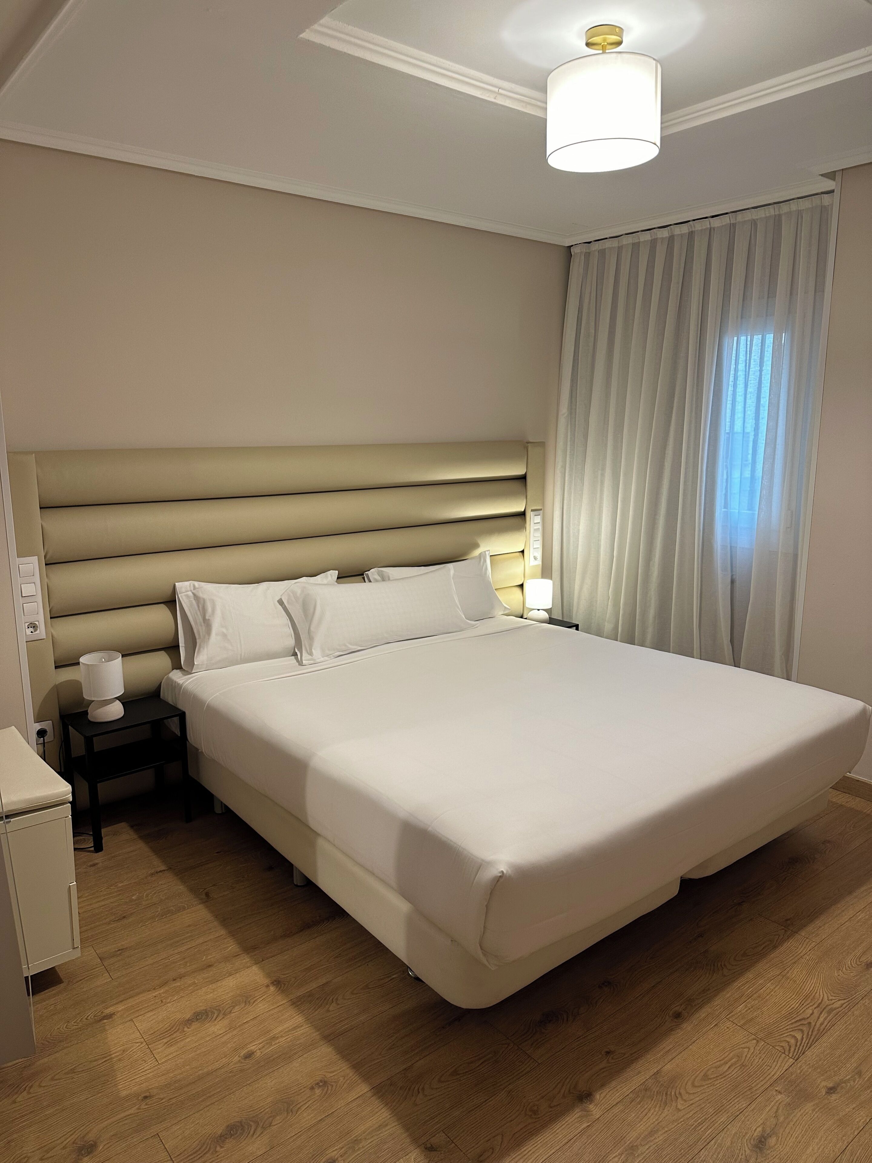 A hotel room with a large bed, a bedside table with a lamp, a nightstand, and a wooden floor.