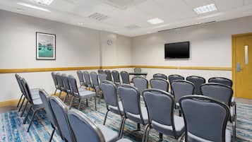 Un salón de conferencias con filas de sillas, un reloj, un televisor de pantalla plana y un cuadro en la pared.