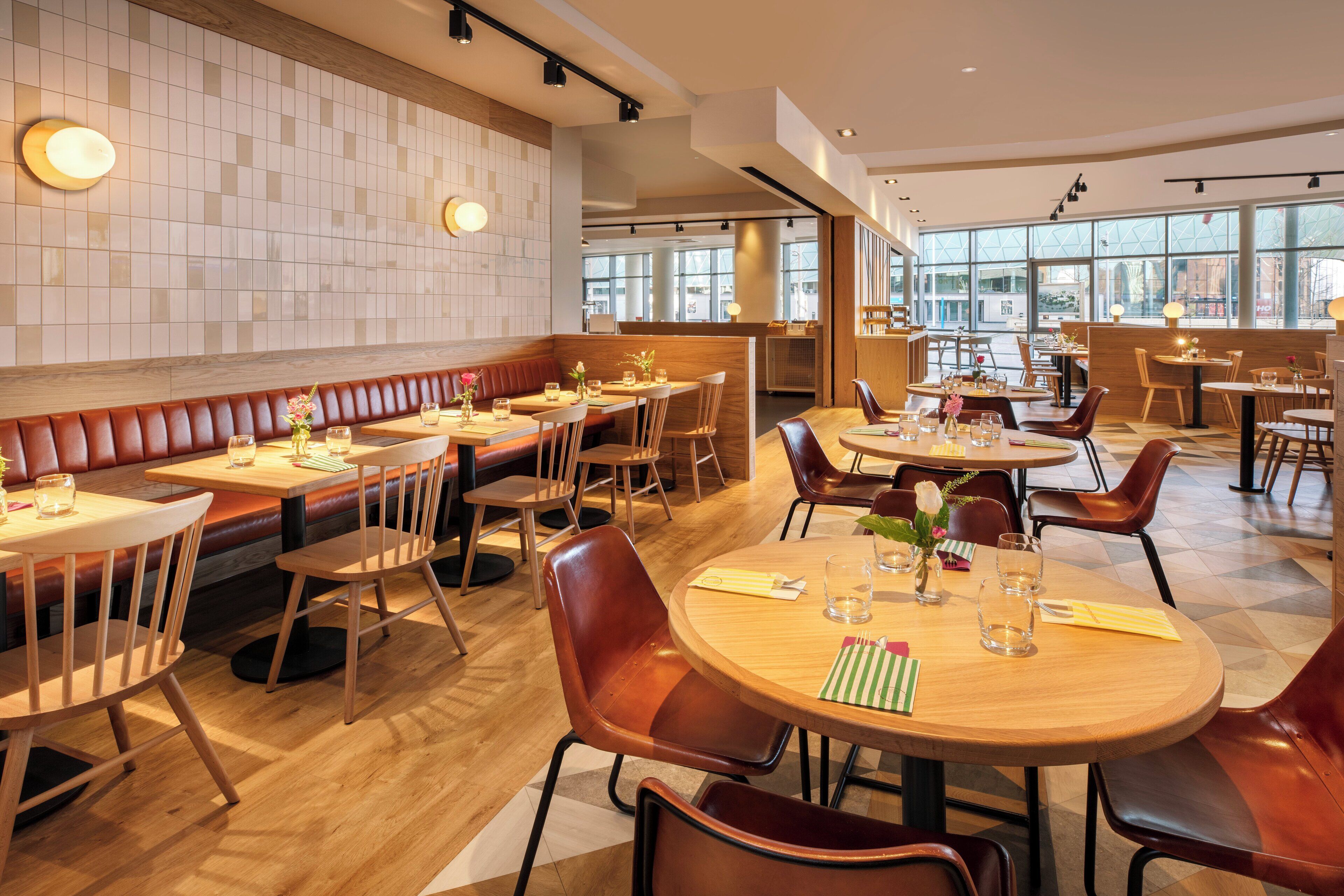 Un ristorante moderno con tavoli e banconi in legno, ampie finestre e una parete rivestita di piastrelle.