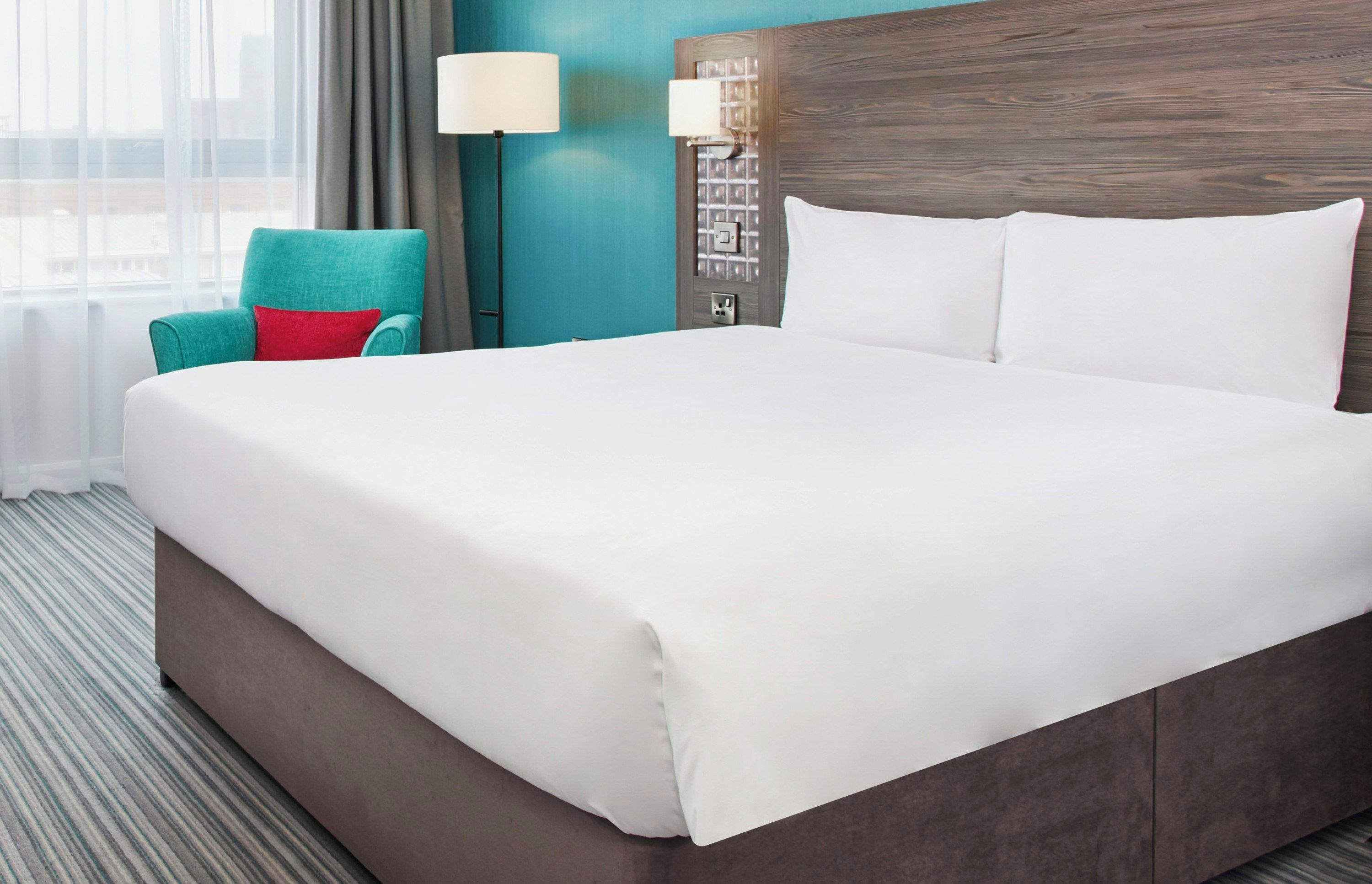 A hotel room with a large bed, a teal armchair, a lamp, and a wall-mounted light fixture.