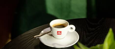 Una taza de café sobre un platito con una cuchara, una taza de latte con un diseño de corazón, y una planta verde sobre una mesa.