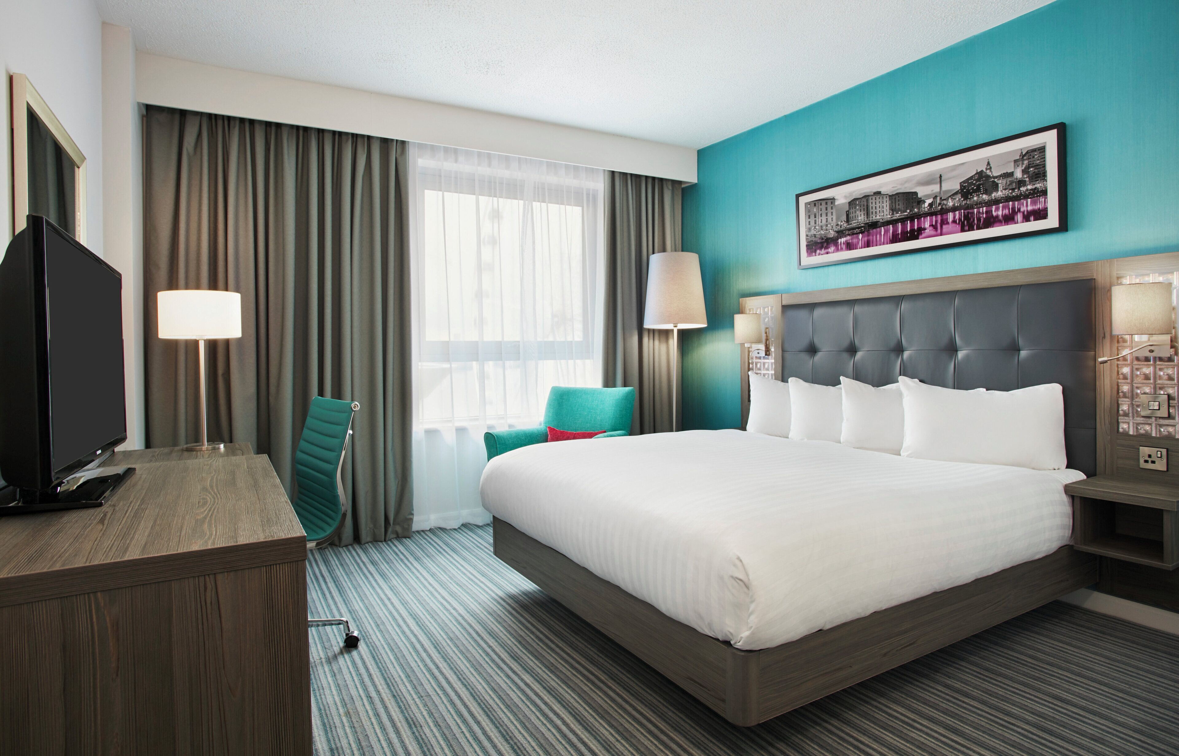 A hotel room with a large bed, a desk with a television, a chair, and a window with curtains.
