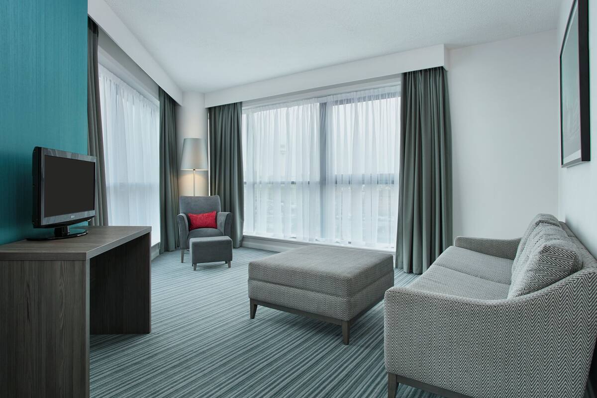 A modern hotel room with a flat-screen TV, a grey armchair with a red pillow, a grey sofa, and a large window with sheer curtains.