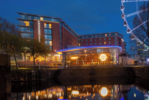 A modern hotel with a prominent curved entrance, a Ferris wheel in the background, and a reflection in the water in front.
