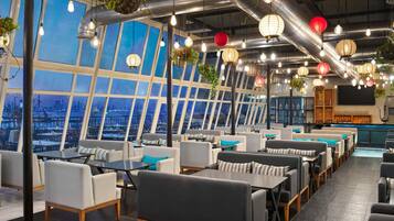 A modern restaurant with high ceilings, exposed ductwork, and large windows offering a city view. Tables and chairs are arranged for dining.