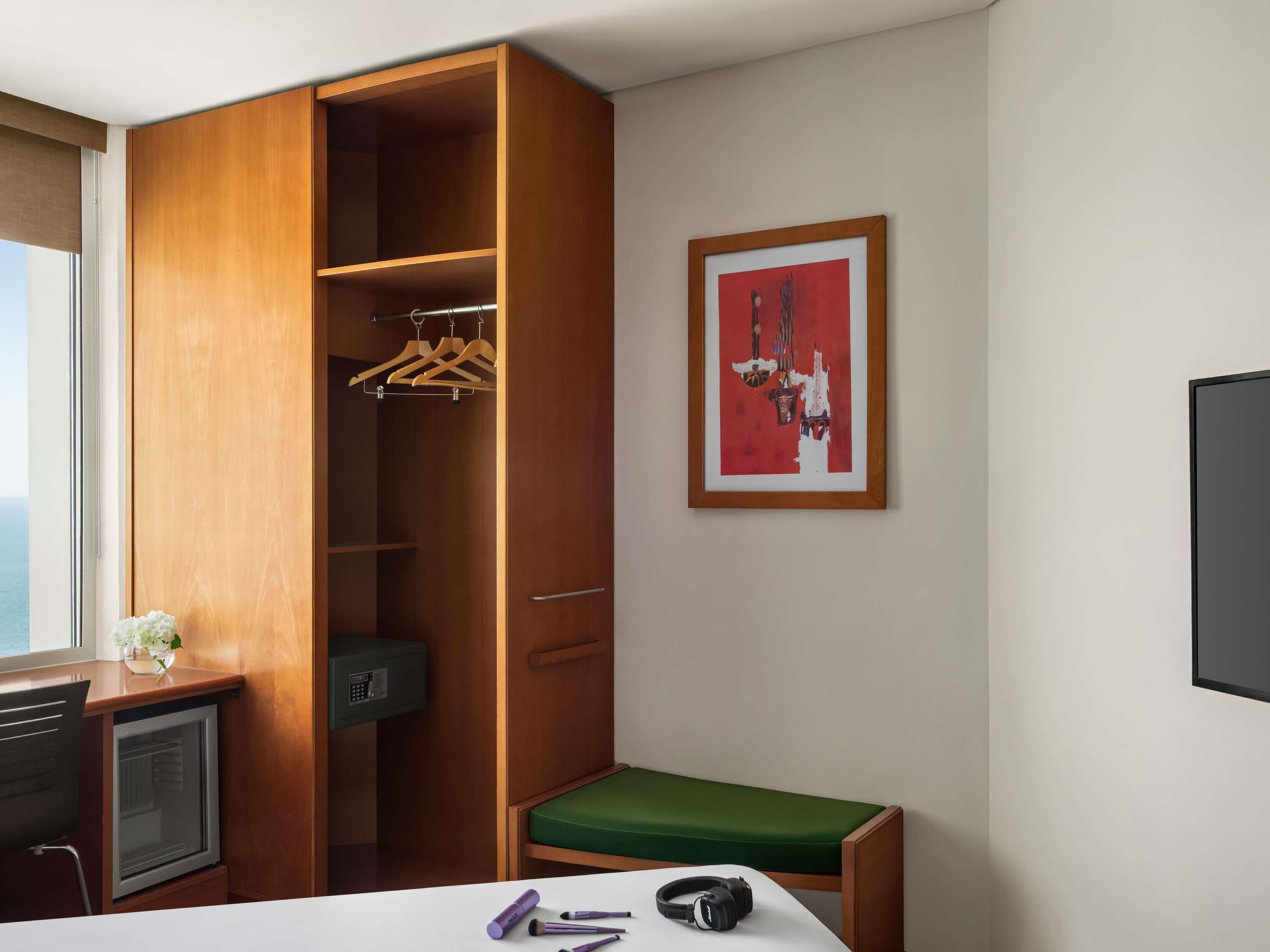 A hotel room with a wooden wardrobe, a desk with a computer, a green cushioned bench, and a framed abstract painting on the wall.