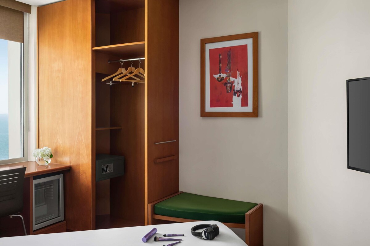 A hotel room with a wooden wardrobe, a desk with a computer, a green cushioned bench, a framed artwork on the wall, and a window with a view.