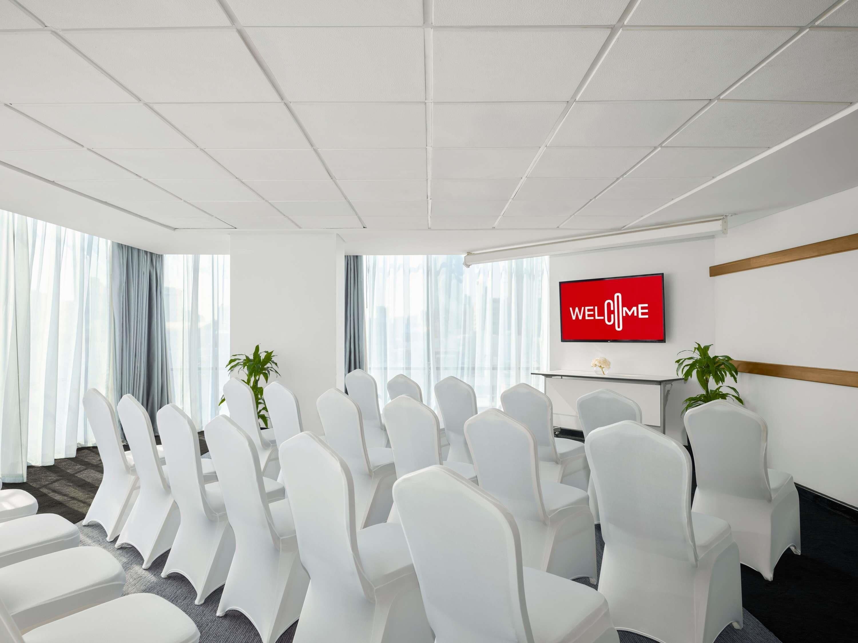 A conference room with white chairs, a TV screen displaying 'WELCOME', and large windows with curtains.
