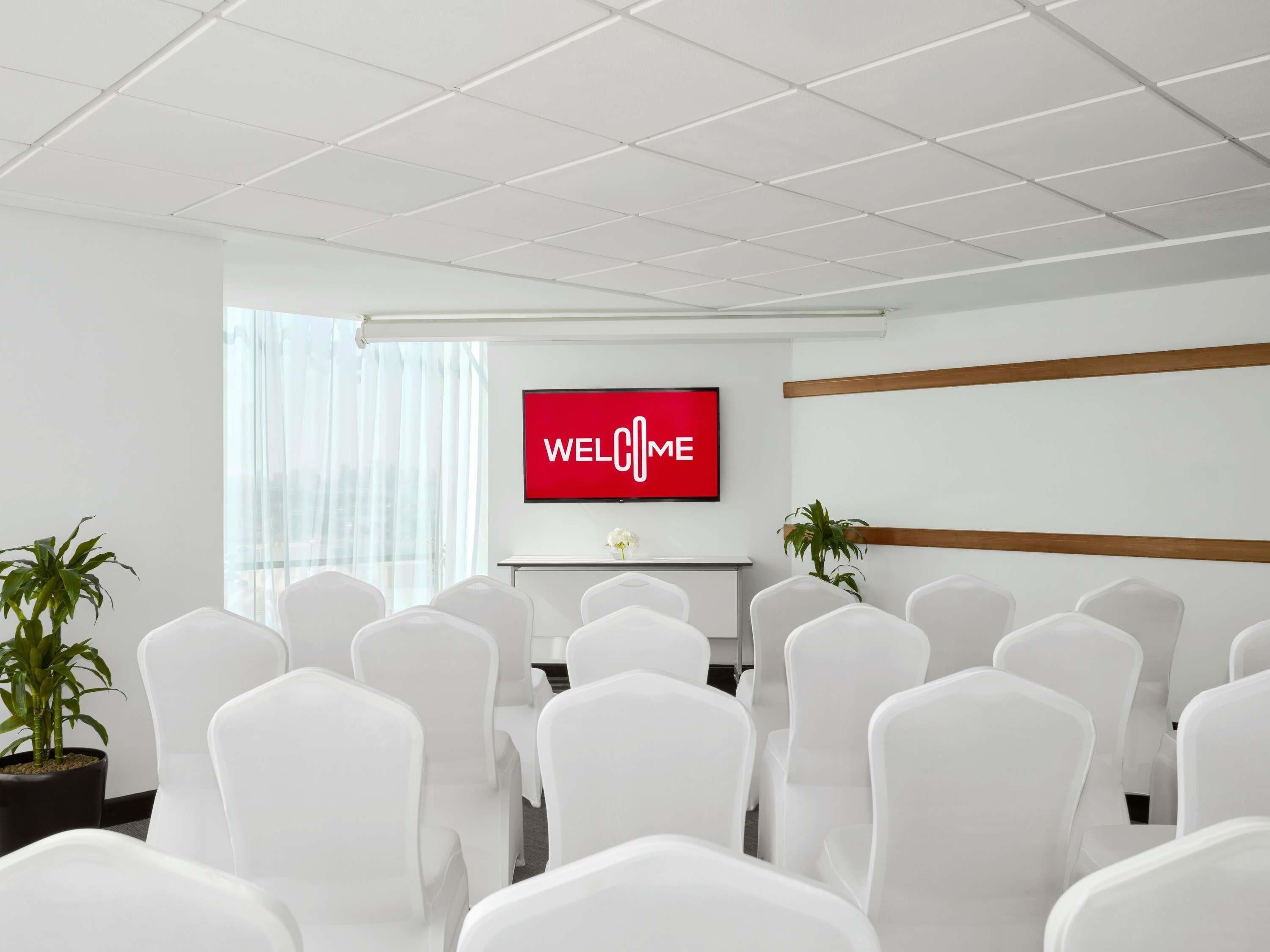 A conference room with white chairs, a 'WELCOME' sign on the wall, a TV, and a window with curtains.