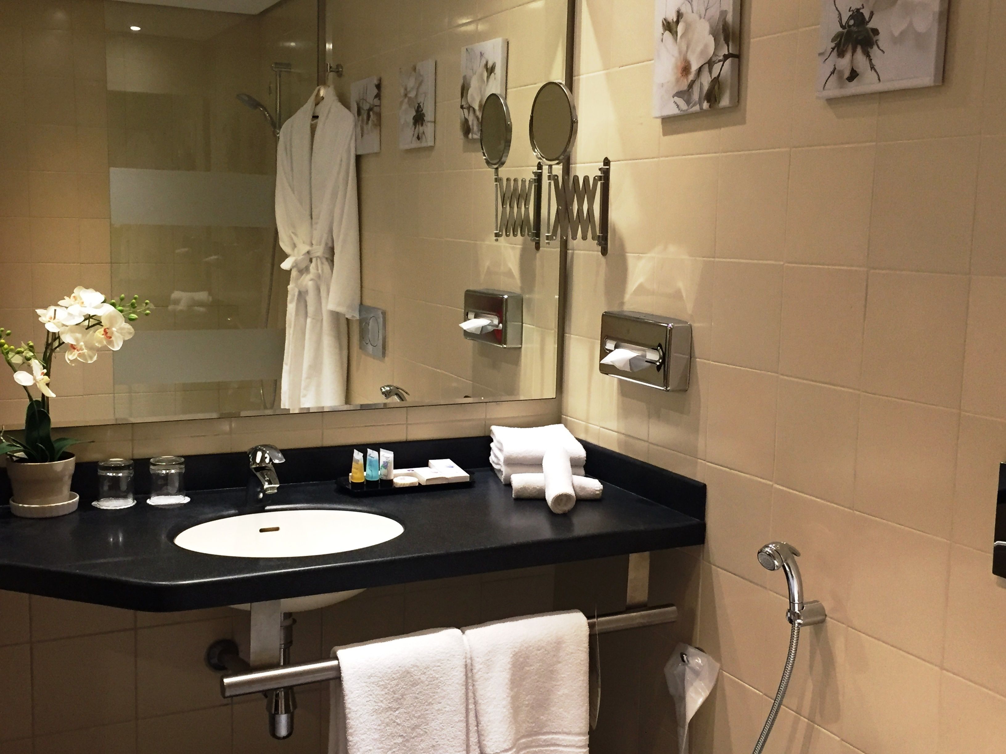 A modern bathroom with a black countertop, white sink, and a large mirror. There are towels and toiletries on the counter, and a shower is visible in the background.
