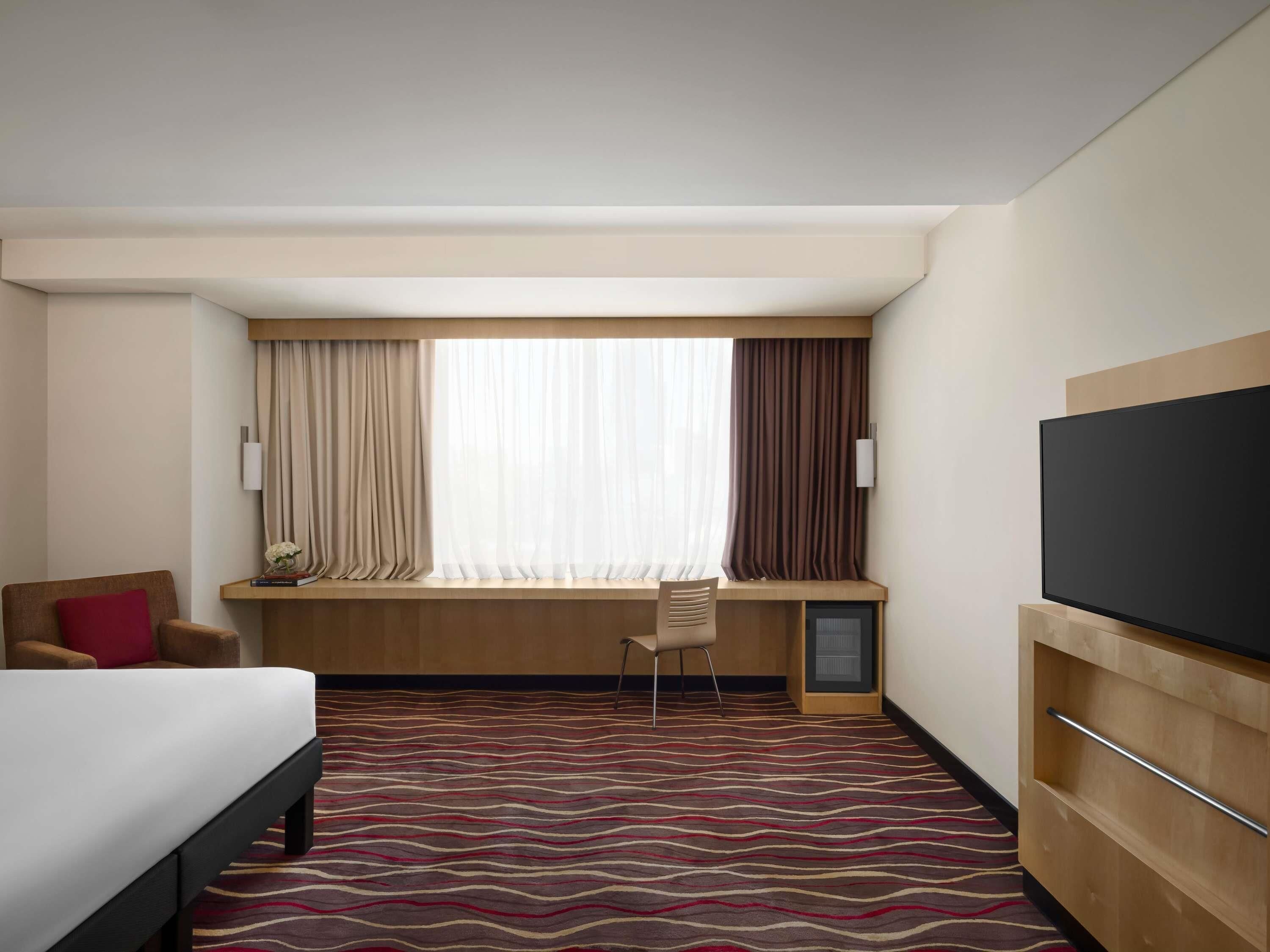 Habitación de hotel con cama, escritorio, silla, televisor y ventana con cortinas.