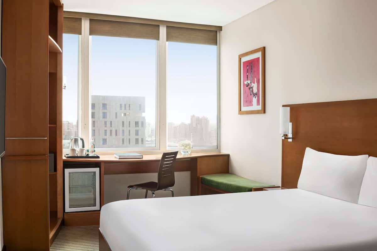 A hotel room with a bed, a desk with a chair, a small table with a kettle, a picture on the wall, and a window with a city view.