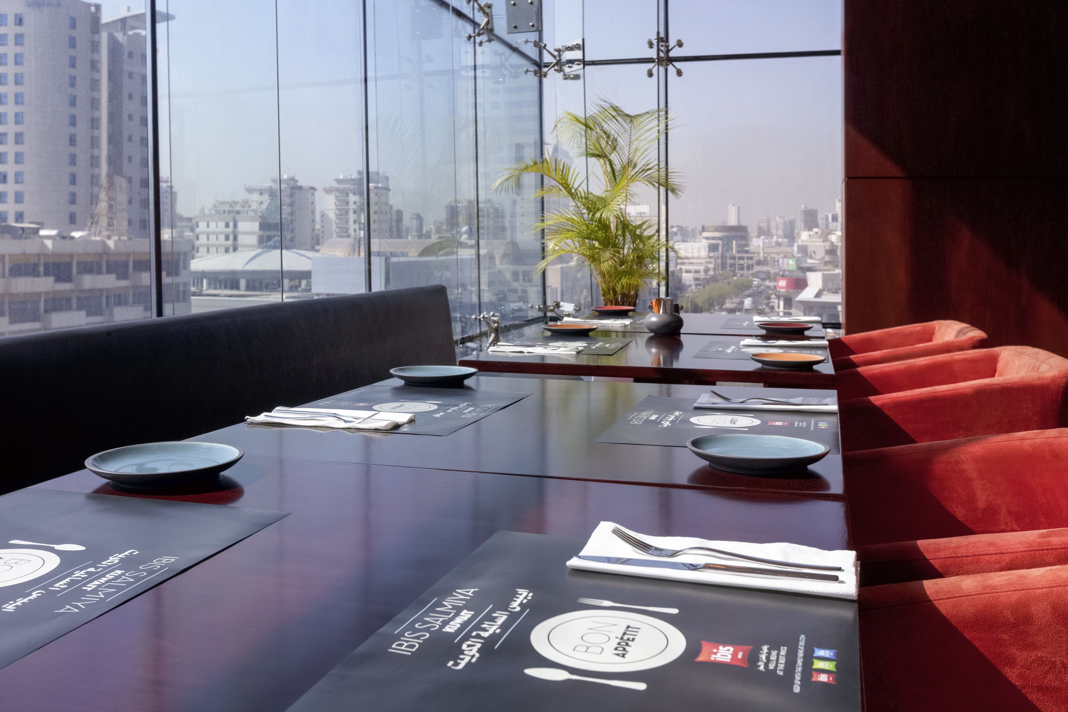 Un restaurante con vista a la ciudad, mesas dispuestas con platos y cubiertos, y una planta en maceta sobre la mesa.