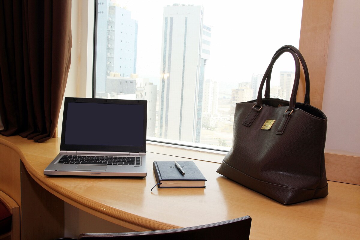 A laptop, a notebook, and a handbag on a desk by a window with a city view.