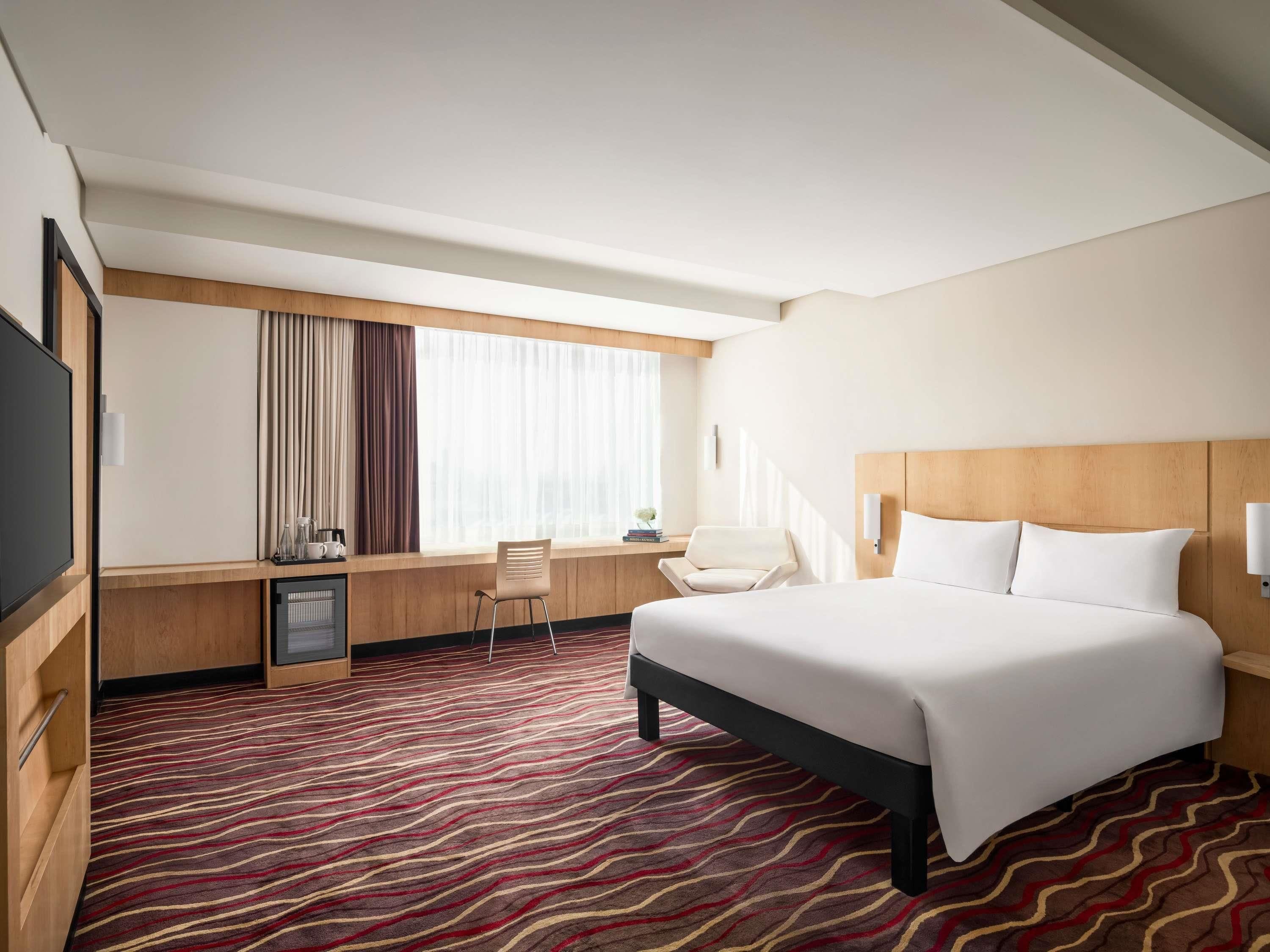 A hotel room with a bed, a desk with a chair, a television, and a window with curtains.
