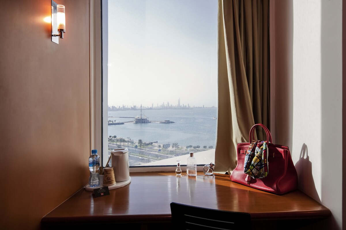 A hotel room with a view of a city skyline and water. There is a table with a water bottle, a candle, and a handbag.