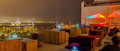 Un bar en la azotea con asientos de madera, luces de neón y vistas del horizonte urbano nocturno.
