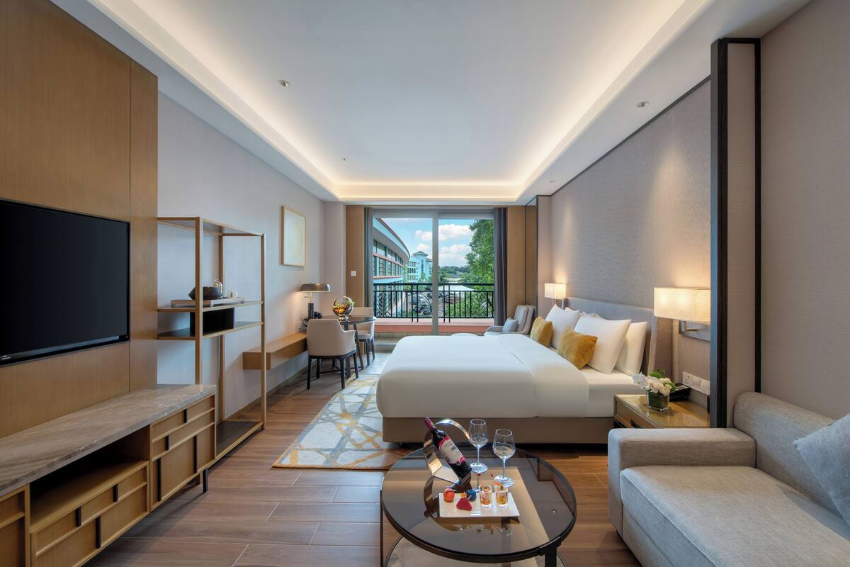 A modern hotel room with a large bed, a TV, a desk, and a balcony with a view.
