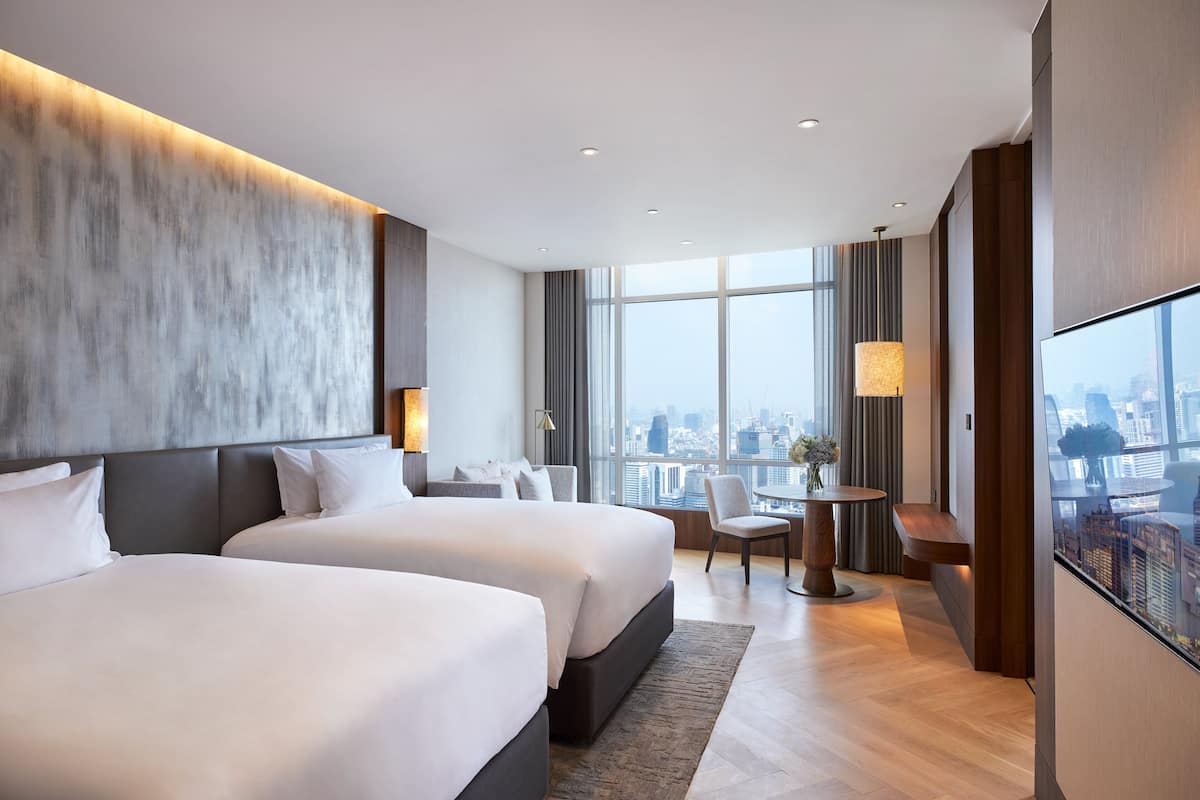 A modern hotel room with two large beds, a city view, a dining area, and a large TV screen.