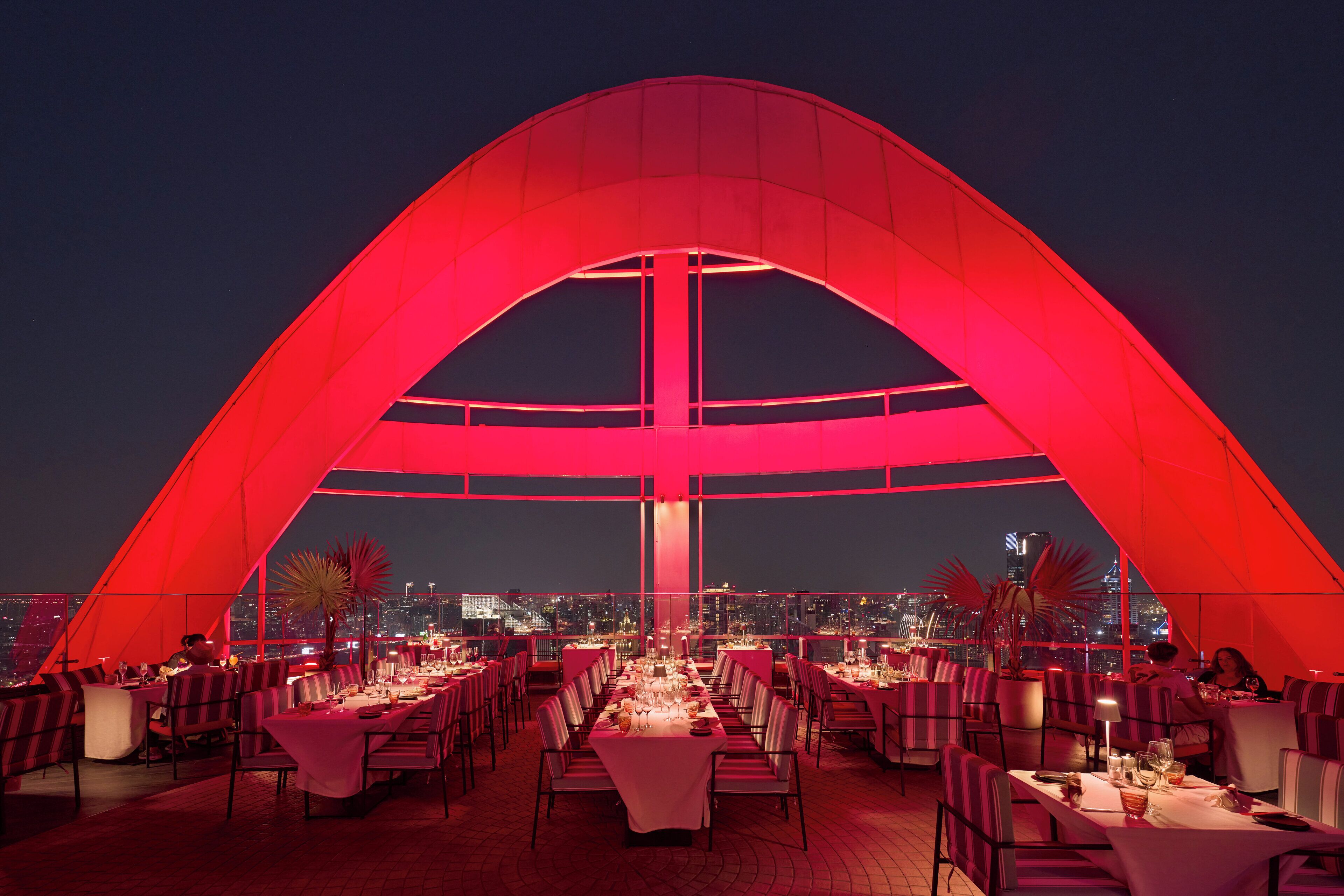 A rooftop dining area with tables set for dinner, a large red arch structure, and a city skyline view in the background.