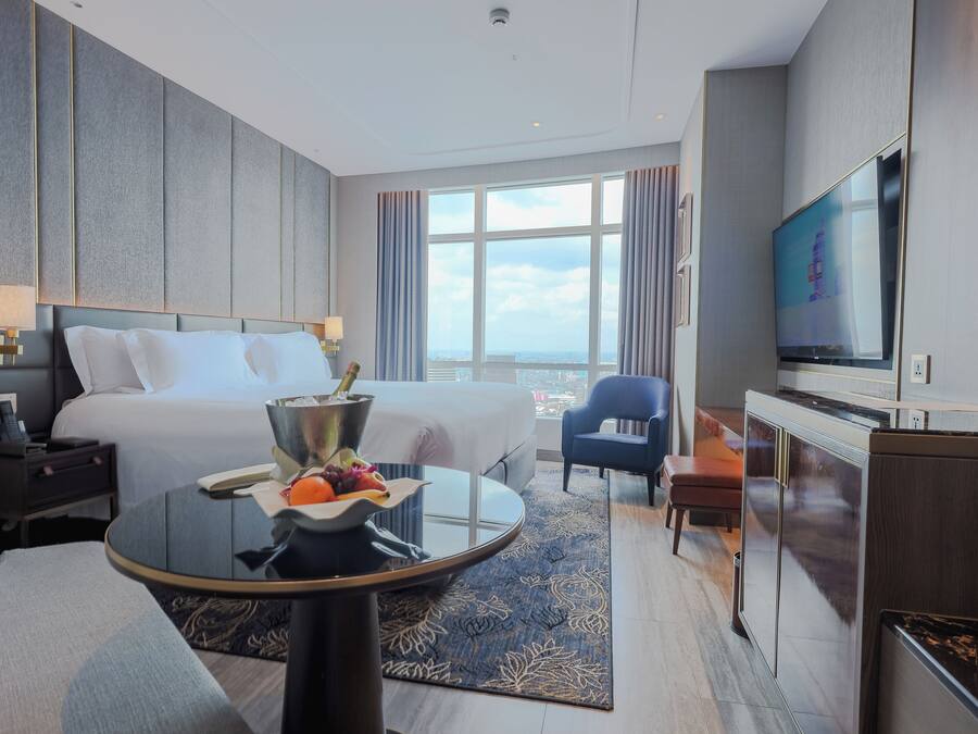 A modern hotel room with a large bed, a TV, a desk, and a view of the city.