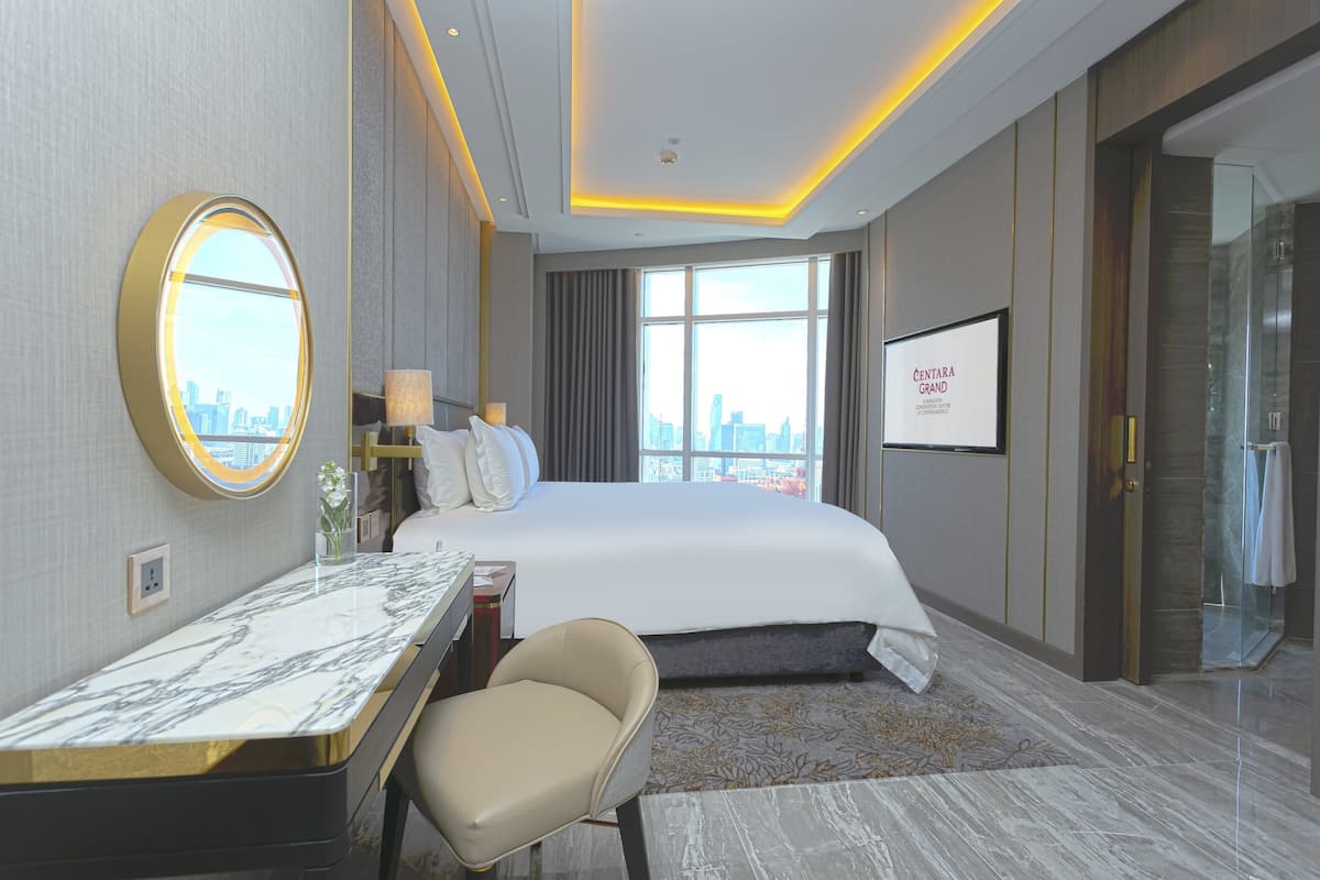 A modern hotel room with a large bed, a desk, a chair, a round mirror, and a city view.