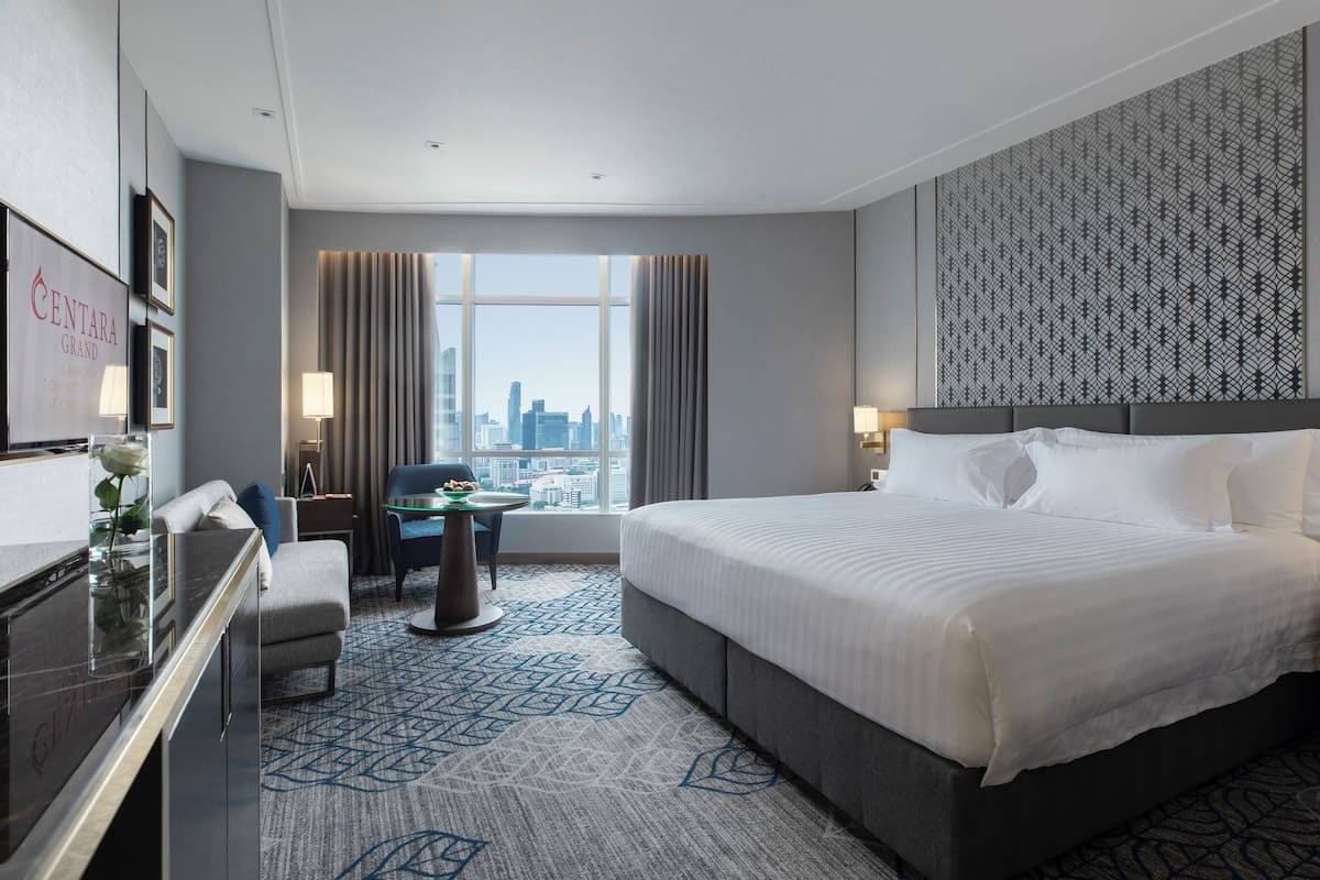 A hotel room with a large bed, a desk, a chair, a view of the city, and a patterned wall.