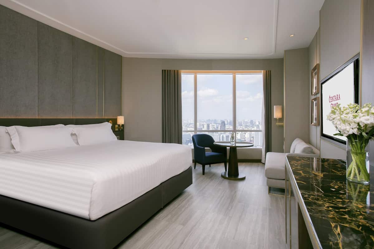 A modern hotel room with a large bed, a small round table, a chair, a sofa, and a large window offering a city view.